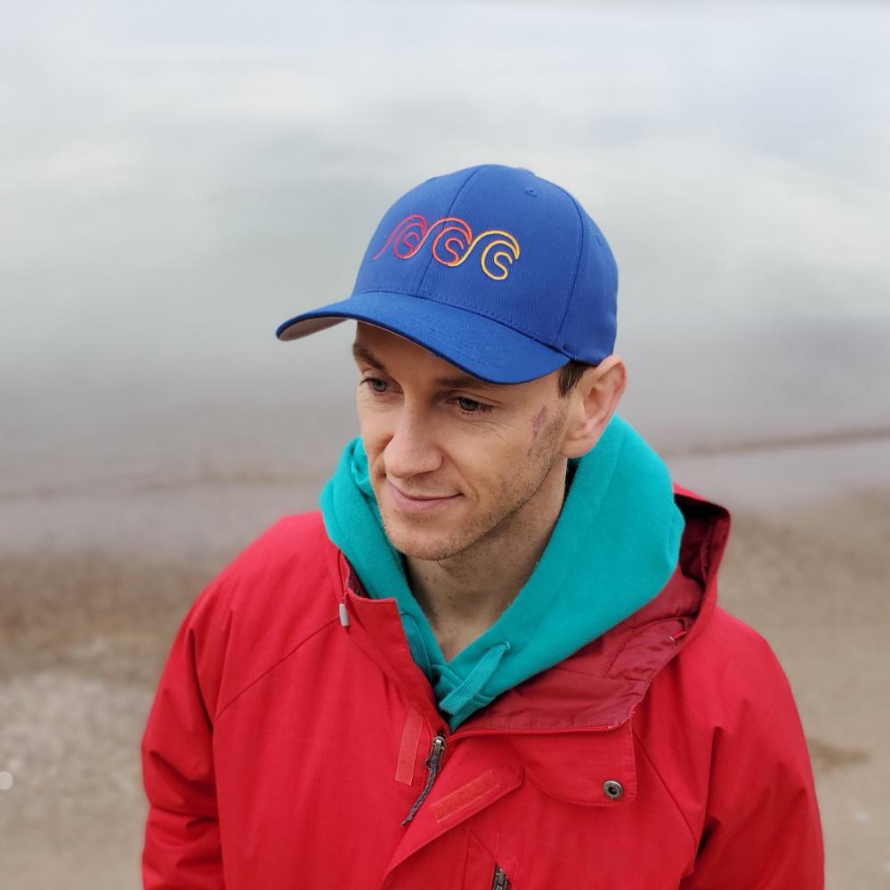 Man with blue hat with a tri logo Calisober Clothing hat wearing a red winter jacket with the subject taking up more than 50% of the screen with a beach in the background.