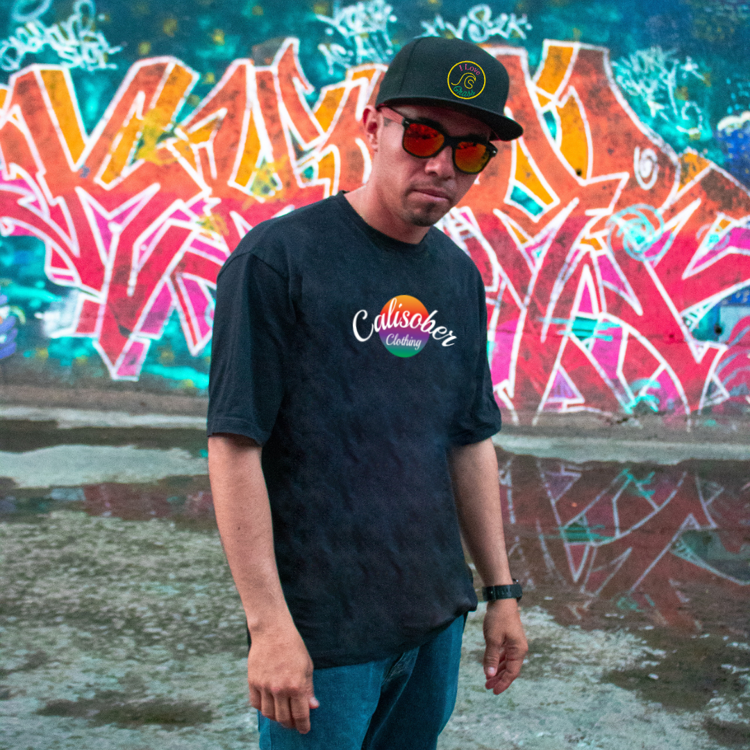 guy with dark skin wearing a black Calisober Clothing t-shirt with an I love grass hat, wearing sunglasses with graffiti in the background. 