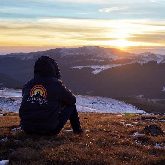 person sitting with back to picture on a hill looking out at the mountains at sunset with some snow on the ground in the distance.
